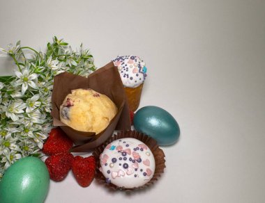 Süslü kapkekler, renkli yumurtalar, kuru çilekler ve açık arkaplanda beyaz bahar çiçekleri olan Paskalya kompozisyonu