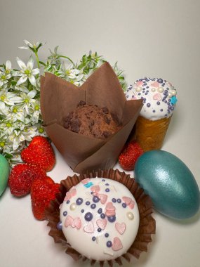 Paskalya çörekleri, çikolatalı kekler, renkli yumurtalar, çilekler ve tarafsız bir arkaplanda bahar çiçekleri.