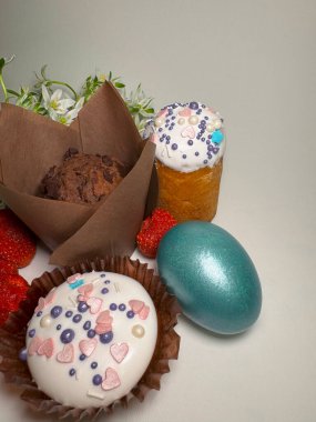 Temiz bir arkaplanda neşeli kapkekler, çikolatalı kek, boyanmış yumurta, çilek ve beyaz bahar çiçekleri ile Paskalya düzenlemesi.