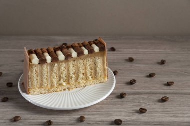 Beyaz bir tabakta servis edilen çikolata ve krema sosuyla süslenmiş katmanlı vanilyalı ve kremalı bir kekin yakın çekimi. Kahve çekirdekleri ahşap bir yüzeye saçılmış, samimi ve davetkar bir tatlı kompozisyonu yaratıyor.