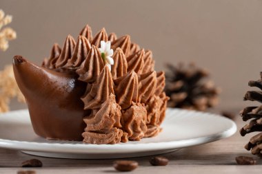 Kirpi şeklinde çikolatalı bir tatlıya yakın plan beyaz bir tabakta krema ile çevrili, çam kozalakları ve kahve çekirdekleri ile çevrili, sıcacık, kırsal bir ruh hali uyandıran