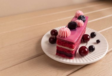 Üzerinde pembe krema, kiraz ve yaban mersini olan lezzetli bir dilim çilekli pasta, ahşap bir masada beyaz bir tabakta servis edilir. Tatlı menüleri, yemek blogları ve tatlı tarif çizimleri için mükemmel.