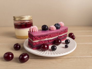 Beyaz bir tabakta ustalıkla sunulmuş pembe kremalı ve taze meyveli bir dilim çilekli pasta. Arka planda, bir kavanoz katmanlı tatlı, bu fotoğrafı yemek blogları ve yemek tarifi web siteleri için mükemmel kılıyor.