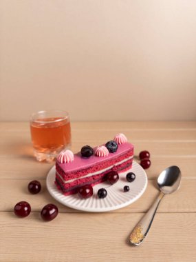 Beyaz bir tabakta servis edilen, yaban mersini ve kirazla süslenmiş pembe kremalı bir dilim pasta. Yanında da bir bardak çay ve ahşap bir yüzeyde dekoratif bir kaşık.