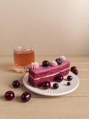 Bir dilim katmanlı kek pembe kremalı, yaban mersini ve kirazla süslenmiş, yanında da bir bardak çay. Tatlı beyaz bir tabakta, etrafa dağılmış böğürtlenlerle hazırlandı.