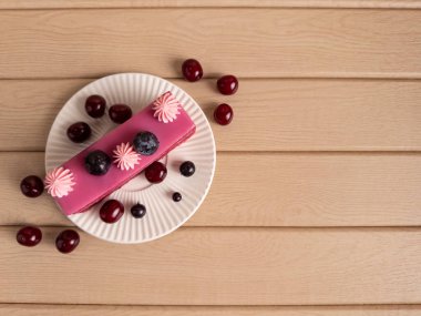 Üzerinde pembe şekerleme çiçekleri, yabanmersinleri ve kirazlar olan parlak pembe jöleli dikdörtgen pasta. Kırsal ahşap bir yüzeyde beyaz bir tabakta sunulan zarafet ve doğal cazibenin bir karışımı.