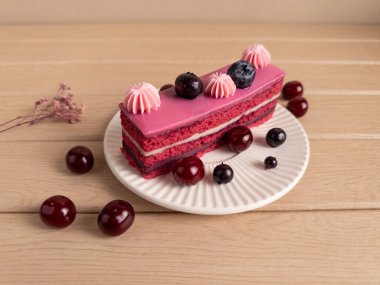 Bir dilim çilekli pasta pembe kremalı, kirazlı, yaban mersinli ve beyaz tabakta kremalı. Ekstra böğürtlen ve kurumuş çiçekler, hafif ahşap bir yüzeyde. Kırsal ve iştah açıcı bir dokunuş ekleyin.