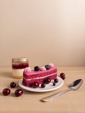 Üzerinde yaban mersini ve kiraz olan pembe kremalı bir dilim pasta. Kavanozlu tatlı ve gümüş kaşığın yanında. Bej arka planı olan ahşap bir yüzeye kurulmuş. Gıda içeriği için mükemmel