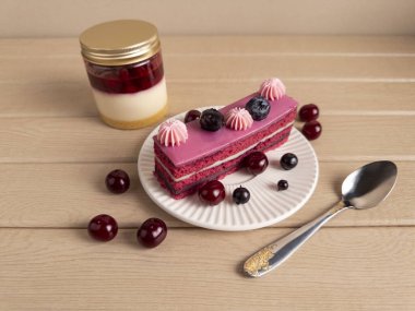 Bir dilim çilekli pasta pembe kremalı ve beyaz tabakta taze böğürtlenli, katmanlı kavanoz tatlısının yanında ve altın detay içeren gümüş bir kaşık. Samimi ve davetkar bir görünüş için hafif ahşap bir yüzey ayarlayın.