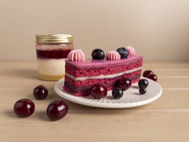 Bir dilim kırmızı kadife pasta pembe kremalı ve bir kavanoz katmanlı tatlının yanında böğürtlenli. Parlak renkler ve düzgün bir düzen onu tatlı temalı ve mutfak içeriği için ideal hale getirir.