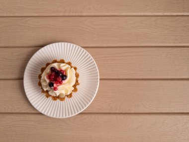 Krem şanti ve taze böğürtlenle kaplı mini tart, tahta bir arka planda beyaz bir tabağa yerleştirilmiş. Fırın promosyonları, tatlı menüleri ve yemek blogları için ideal.