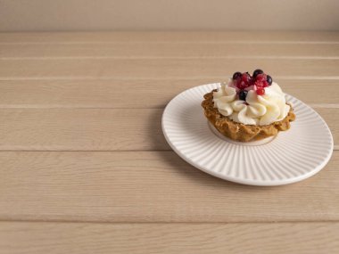 Beyaz kremayla kaplanmış, beyaz yaban mersini ve kırmızı frenk üzümlü, beyaz bir tabağa beyaz ahşap bir yüzey üzerine yerleştirilmiş, kahverengi kabuklu, nefis bir turtanın yakın plan görüntüsü.