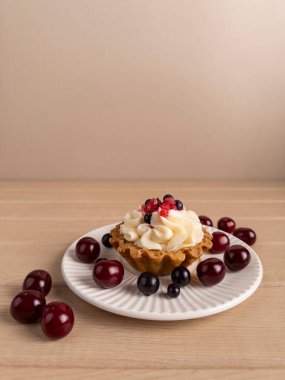 Krem şanti ve böğürtlenle kaplı, beyaz bir tabakta vişne ve kuşüzümü ile çevrili bir kekin yakın çekimi. Asgari ahşap arka planda yakalanmış, fırın promosyonları, yemek blogları ve tarif siteleri için ideal