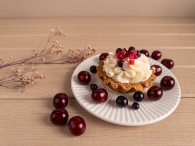 Krem şanti ve taze böğürtlenlerle kaplı, beyaz bir tabakta vişne ve kuşüzümü ile çevrili bir kapkekin yakın çekimi. Kurumuş bir çiçek filizi ahşap arka plana kırsal bir dokunuş ekler.