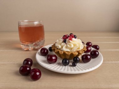 Krem şantili, kırmızı üzümlü ve beyaz tabakta yaban mersinli meyveli tart. Etrafı kirazlarla çevrili ve bir bardak kırmızı meyve suyu açık tahta bir yüzeyde. Yemek temalı görseller için mükemmel.