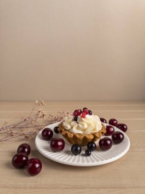 Krem şantili bir tart ve beyaz bir tabakta vişne ve yabanmersini de var. Daha fazla böğürtlen ve kurumuş çiçeklerle çevrili, açık ahşap bir yüzeye kurulmuş.