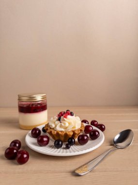 Kirazlar ve katmanlı tatlı kavanozuyla tamamlanmış taze dutlu çırpılmış kremalı tart içeren göz kamaştırıcı bir tatlı düzeneği. Hepsi de açık ahşap bir yüzeye ustalıkla yerleştirilmiş.