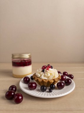 Beyaz bir tabakta çırpılmış krema ve meyveli küçük bir tart. Etrafı kirazlarla çevrili. Arka planda bir kavanoz meyveli, kremalı ve pastalı tatlı var. Yemek blogları ve mutfak temaları için ideal