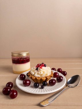 Krem şantili tart ve kirazlı bir tabakta, kavanozda katmanlı bir tatlının yanında. Ahşap masada rahat, dikey yemek kompozisyonu.