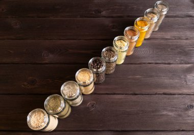 Cam kavanozlarda düzgünce düzenlenmiş çeşitli baharat ve baharat koleksiyonu. Kavanozlar, koyu arkaplana karşı ahşap bir yüzeye yerleştiriliyor ve içeriğinin rengini ve dokusunu vurguluyor.