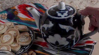 Geleneksel bir Özbek çaydanlığından bir el renkli atlas kumaş üzerine küçük bir çay fincanına çay dolduruyor, yanında da bir tabak halva var.