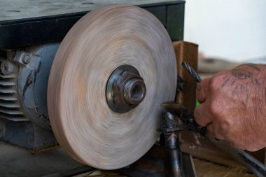 Bir atölyede dönen çarkın üzerinde metal bir aleti öğüten bir işçinin yakın çekimi. Kıvılcımlar uçar, bu endüstriyel anda işçiliğin makineyle buluşması gibi.