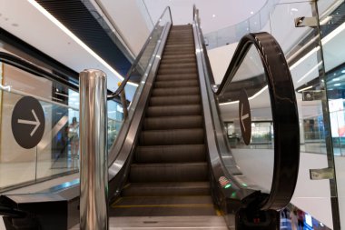 Çağdaş kapalı alışveriş merkezleri, cam parmaklıklar, fayanslı döşemeler, markalı mağazalar, ve merkezi yürüyen merdivenlere yakın insanlar.