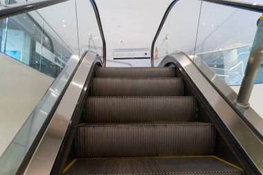 Çağdaş kapalı alışveriş merkezleri, cam parmaklıklar, fayanslı döşemeler, markalı mağazalar, ve merkezi yürüyen merdivenlere yakın insanlar.