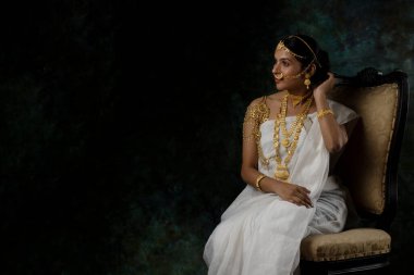 Tek başına, beyaz sari giyen, altın takılar takan, yeşil, saf bir arka planda altın bilezik takan Hintli bir kadın resmi.