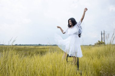 Siyah saçlı, buğday tenli, beyaz etekli ve açık çim tarlasının ortasında poz veren genç bir Hintli kadın model.