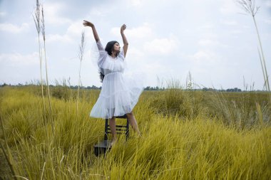Siyah saçlı, buğday tenli, beyaz etekli ve açık çim tarlasının ortasında poz veren genç bir Hintli kadın model.