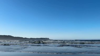 Dalgalar, arka planda daha büyük dalgaların çarptığı kumlu bir sahile doğru kırılıyor. Mavi gökyüzü ve açık bir gün. Tofino yakınlarında, İngiliz Kolombiyası