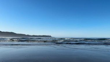 Dalgalar, arka planda daha büyük dalgaların çarptığı kumlu bir sahile doğru kırılıyor. Mavi gökyüzü ve açık bir gün. Tofino yakınlarında, İngiliz Kolombiyası