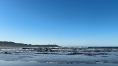 Dalgalar, arka planda daha büyük dalgaların çarptığı kumlu bir sahile doğru kırılıyor. Mavi gökyüzü ve açık bir gün. Tofino yakınlarında, İngiliz Kolombiyası