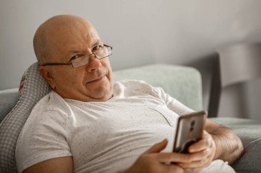 Elinde cep telefonu olan gözlüklü yetişkin bir adam koltukta rahatça oturuyor.