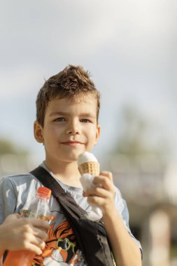 Kahverengi gözlü esmer çocuk, 6-7 yaşlarında, güneşli bir günde parktaki gözleme külahında dondurma yiyor.