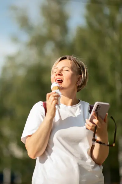 an adult blonde in a white T-shirt with a mobile phone in her hands, happily eating ice cream in a waffle cone in the park on a sunny day