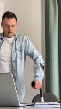 Koltuk değnekleriyle yürüyen bir adam evden çalışmak ya da internet randevusu için dizüstü bilgisayarın başına oturur. Koltuk değneği engelli. Dikey 4K beyaz. Yüksek kaliteli FullHD görüntüler