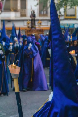 Andalusia, Spain. Nazarene capiruchos in spanish Holly Week procession holding candles to illuminate the path for Jesus Nazareno, in bokeh background. Purple hood, white gloves, vertical shot clipart