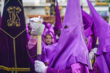April 7th 2023 - Dona Mencia, Cordoba, Spain. Nazarene capirucho devotee fires candle in spanish Holly Week procession in windy day. Purple hood, white gloves, unrecognized person clipart