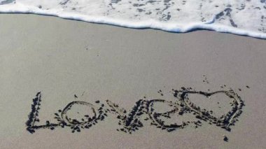 Deniz kıyısındaki kumsalda çizilmiş kalp ve sözcük aşkı. Dalgaların manzarası. Aşkın sembolü. Aşk, Yazılmış ve Çizilmiş Kalp Güzel bir kumsalda Mavi Deniz ve Gökyüzünün Arkaplanına Karşı.