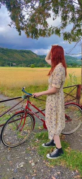 A Young Girl with a Bicycle on the Background of a Field in Italy Outside the City. Landscape With a Bicycle Against the Background of a Field, Road, Green Grass, Summer. Sports and Travel.