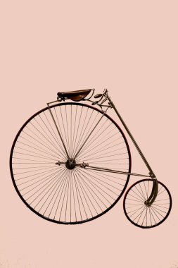 Klasik bir yüksek tekerlekli bisiklet, yumuşak pembe bir arka fona karşı büyük bir ön tekerlek ve daha küçük bir arka tekerleğe sahiptir. Sanatçılık, eşsiz tasarımını ve tarihsel önemini vurguluyor..