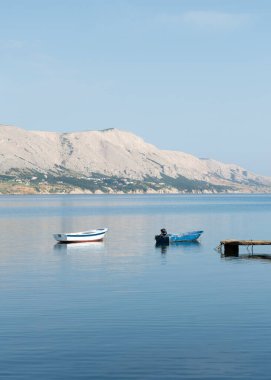 Limana yakın denize demirlemiş iki tekne, arka planda dağlık bir ada.