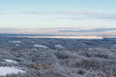 Kırsal kesimin rüzgarlı manzarası, karla kaplı ormanlar ekilebilir araziler, puslu uzak tepeler.