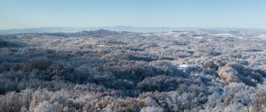 Soğuk kış gecesinden sonra buzla kaplı tepe köyü, evlerle kaplı kırsal alan, tepenin tepesinde yaprak döken orman ve kilise, dağ sıraları.