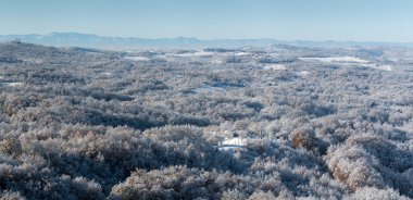 Soğuk kış gecesinden sonra buzla kaplı tepe köyü, evlerle kaplı kırsal alan, tepenin tepesinde yaprak döken orman ve kilise, dağ sıraları.