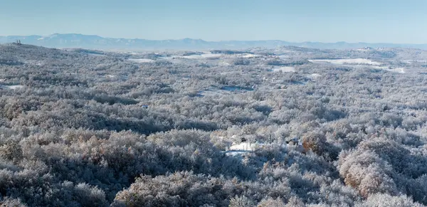 Soğuk kış gecesinden sonra buzla kaplı tepe köyü, evlerle kaplı kırsal alan, tepenin tepesinde yaprak döken orman ve kilise, dağ sıraları.