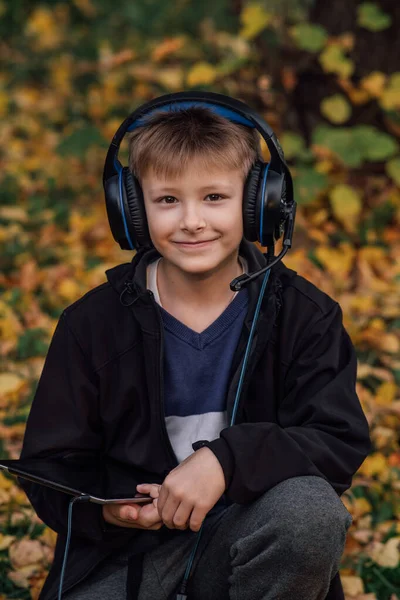Siyah ceketli, okul yakınlarında tablet ve kulaklıklı Avrupalı bir sarışının yakın plan portresi. İnternetten öğrenme amaçlı. Parkta mutlu bir genç adam. Mutlu bir genç. Sonbahar fotoğrafı, ön görünüm