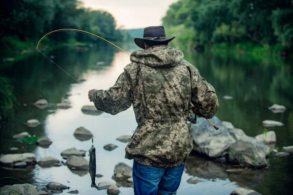 若い男釣りのバックビュー ロッドを持つ漁師 川の岸にリールを回転させます 湖で釣りをしながらロッドを引く魚を捕まえる男 野生の自然 農村部の休暇の概念 — ストック写真
